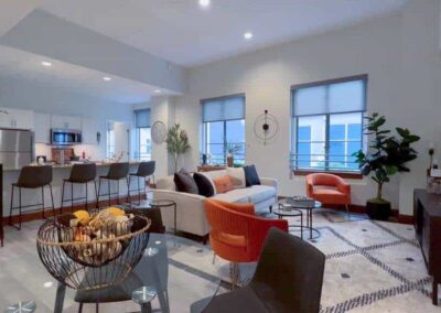 A spacious and modern open-concept living room and kitchen area. The living room features sofas, a pair of orange accent chairs, a round coffee table, and large windows with white blinds. The kitchen has a breakfast bar with high stools, white cabinetry, and stainless steel appliances.