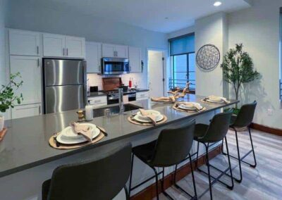 Modern kitchen with a sleek gray countertop and island, around which are four black bar stools. The island is set with plates and napkins. In the background, white cabinets, stainless steel appliances, and a plant on the left. Wall decorations and large windows present.